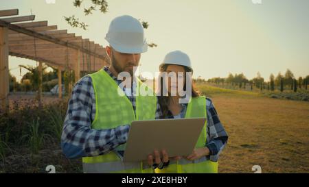 Caucasian colleagues engineers hardhat uniform male man holding laptop pc computer guy speak discussing data talking to female woman girl worker Stock Photo