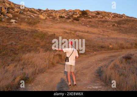 a mature man with binoculars walks along the road in a mountain valley, pointing at the stones with his hand. travel and tourism. Stock Photo