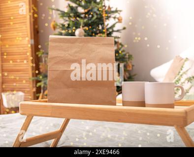 . Paper bag, kraft delivery package, tea cups, mugs on wooden tray on bed, Christmas holiday vibe.. Stock Photo