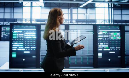 Engineer in server hub checking recovery plan on tablet, monitoring rackmounts energy consumption. Woman in data center making sure rigs are functioning optimally, doing routine disk checks, camera B Stock Photo