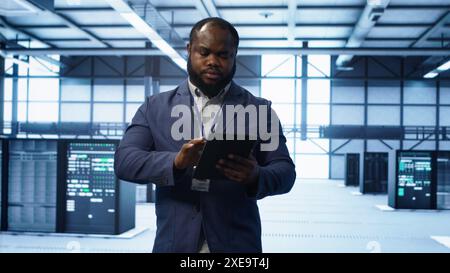 Software developer managing data center with advanced technology, ensuring efficient network management. Admin walking in server room, optimizing data center infrastructure with tablet, camera A Stock Photo