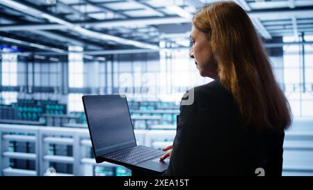 System administrator in data center typing on laptop, running error checking utilities to fix problems. Woman in server hub looking for network issues affecting rigs using notebook device, camera B Stock Photo