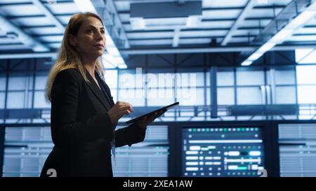 Engineer in server hub checking recovery plan on tablet, monitoring rackmounts energy consumption. Woman in data center making sure rigs are functioning optimally, doing routine disk checks, camera A Stock Photo