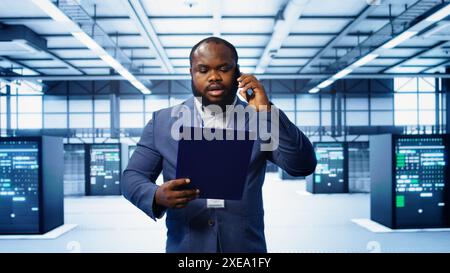 IT professional looking over data center checkup checklist paperwork, talking on phone in server farm. African american man surrounded by server rigs, chatting during telephone call, camera B Stock Photo