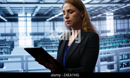 Admin in server room checks recovery plan on tablet, monitoring equipment energy consumption. IT professional uses notebook in data center workplace, ensuring rigs are functioning optimally, camera B Stock Photo