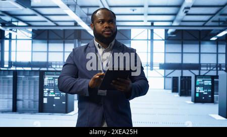 Software developer managing data center with advanced technology, ensuring efficient network management. Admin walking in server room, optimizing data center infrastructure with tablet, camera A Stock Photo