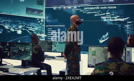 Commander managing cybersecurity measures to protect federal data from enemy breaches, working with his crew from a control center military base. Army officer ensures national safety. Camera B. Stock Photo