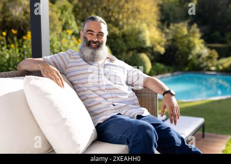 Relaxing by pool, man sitting on outdoor sofa, enjoying sunny day Stock Photo