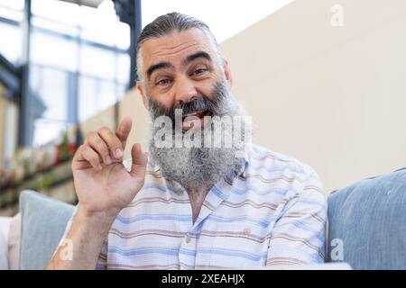 Smiling senior man with beard gesturing while sitting on couch indoors, on video call Stock Photo