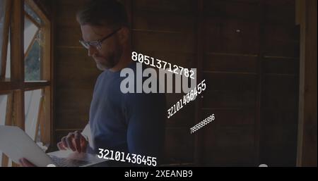 Image of multiple changing numbers over caucasian man standing near window and using laptop Stock Photo