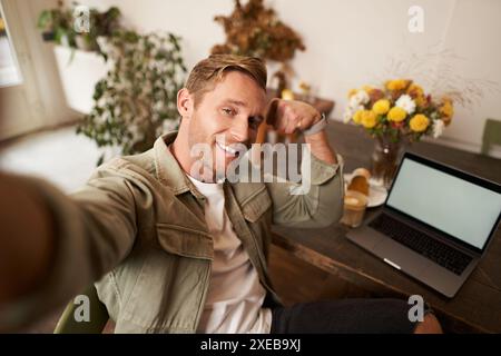 Portrait of handsome and confident man taking a selfie, flexing biceps, shows-off his muscles, sits in cafe in front of laptop, Stock Photo