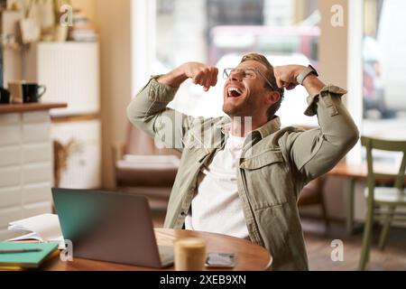 Portrait of pleased, smiling young man shouting with rejoice, sitting in cafe in front of laptop, flexing muscles, screaming fro Stock Photo