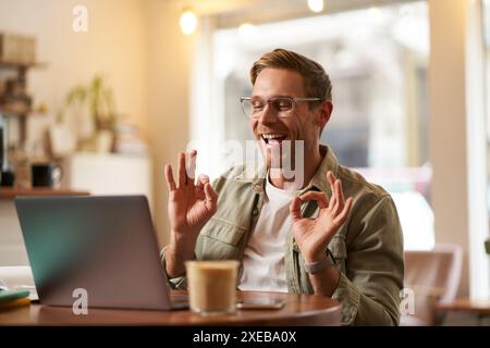 Portrait of young freelancer, digital nomad having an online meeting, video chats, shows okay sign at laptop, confirm something, Stock Photo