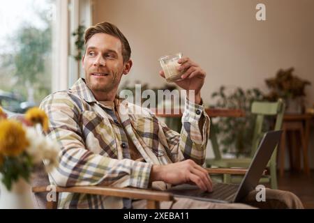Portrait of young freelancer, a man in casual clothes, sitting in cafe and drinking coffee, working remotely on laptop, digital Stock Photo