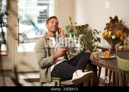 Portrait of handsome happy young man, relaxing in a coffee shop, spending time in cafe, listening to music in wireless headphone Stock Photo