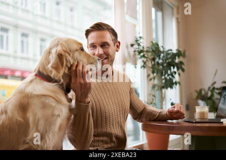 Lifestyle photo of young handsome man, entrepreneur working in a cafe on laptop while his dog sits next to him. A guy pets his g Stock Photo