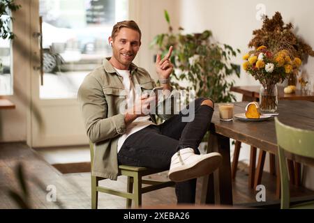 Portrait of happy handsome young man, sitting in cafe with mobile phone, wearing wireless headphones, listening to music or podc Stock Photo