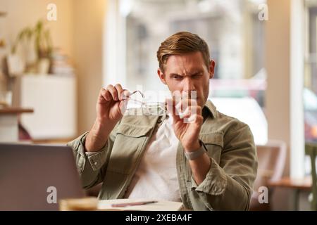 Portrait of man with new anti blue light glasses, looking through the lense at his computer screen, sitting in cafe with laptop Stock Photo