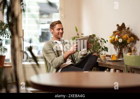 Portrait of handsome happy guy, young man sits in cafe, watching videos on digital tablet, wearing wireless headphones, laughing Stock Photo