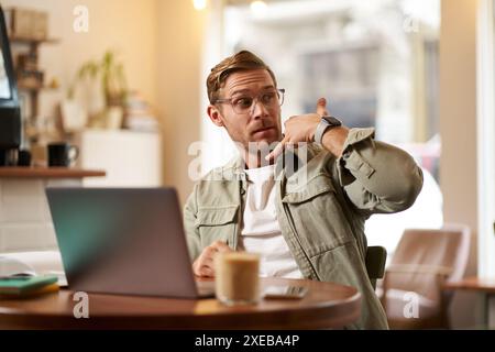 Portrait of young handsome man in glasses, businessman working in cafe, sitting with laptop, showing phone call hand gesture, ad Stock Photo
