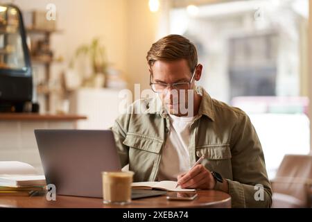Image of focused young man in glasses, sitting in cafe, making notes, studying, attending online course, learning on remote from Stock Photo