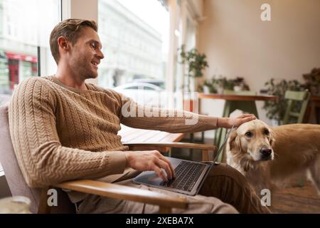 Portrait of young man, digital nomad, sitting in cafe near window, using laptop, working with his pet, petting golden retriever Stock Photo