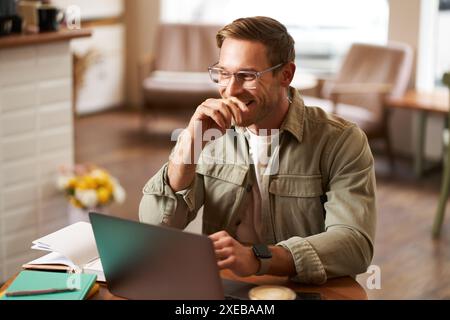 Image of young digital nomad, man in glasses sits in cafe, works from coffee shop, uses laptop in co-working space, wears glasse Stock Photo