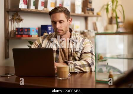 Portrait of young man looking concentrated, working on laptop, freelancer doing his project in cafe, typing on keyboard, drinkin Stock Photo
