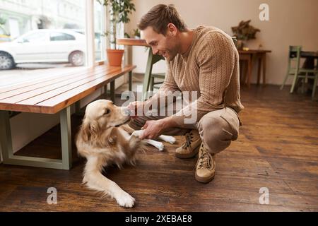Portrait of handsome man playing with his dog in a cafe, a guy holds his pet paw in hand and smiles, spends time in pet friendly Stock Photo