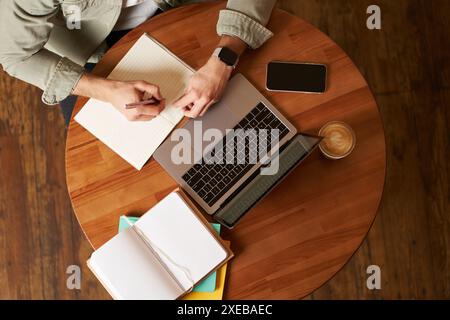 Top aerial view of male hands taking notes, writing in notebook, man sitting at round desk in cafe, working on laptop, studying Stock Photo