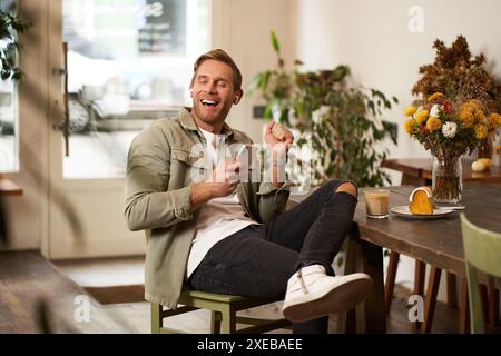 Happy young man in coffee shop, drinking cappuccino, listening to funny podcast in earphones, holding smartphone. Handsome male Stock Photo