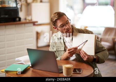 Portrait of handsome young man giving lessons online, showing exercises in his notebook, looking at laptop, recording video, sit Stock Photo