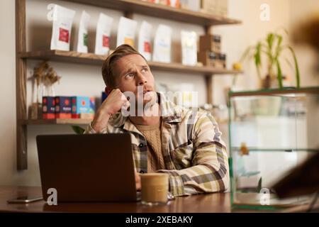 Man looking concentrated while working remotely from a coffee shop. Business owner sitting in cafe with laptop, drinking cappucc Stock Photo