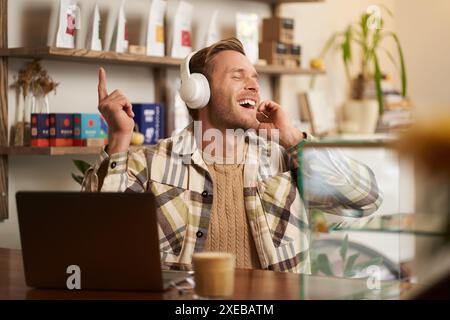 Portrait of handsome happy guy, freelancer working and listening to music in wireless headphones, dancing on his chair in cafe, Stock Photo