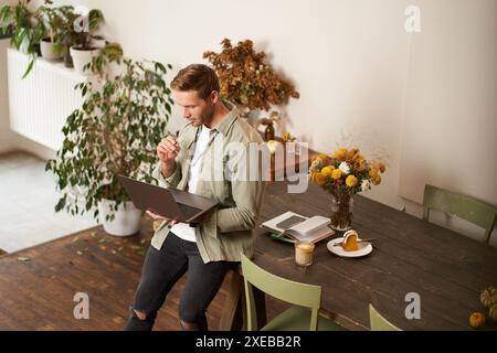 Portrait of young manager, man sits on table, holds laptop, looks concentrated at screen, reading project information, checking Stock Photo