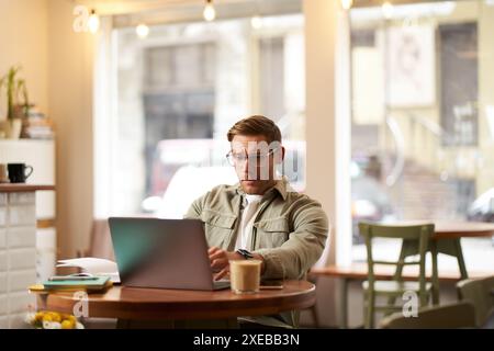 Image of young digital nomad, man in glasses sits in cafe, works from coffee shop, uses laptop in co-working space, wears glasse Stock Photo
