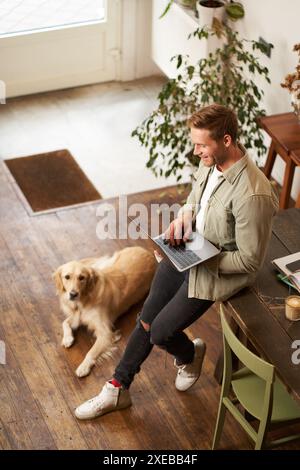 Vertical shot of handsome businessman, young man with laptop leans on table, sitting at co-working space with his dog. Young sal Stock Photo