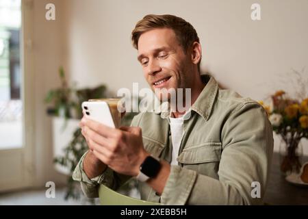 Image of handsome young man in cafe, looking at his mobile phone, using smartphone app, drinking coffee Stock Photo
