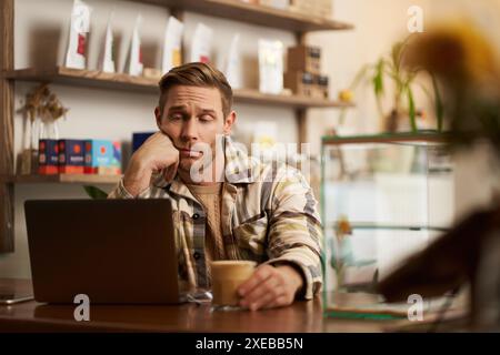 Portrait of man looking disappointed at laptop screen, working remotely, freelancer sitting in cafe, drinking coffee, reading me Stock Photo