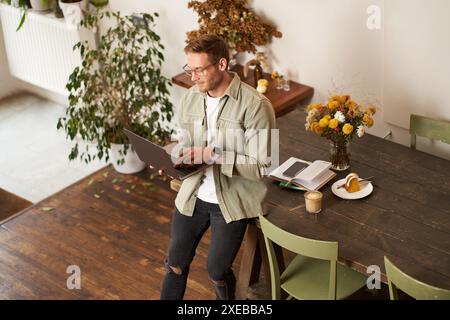 Handsome manager, man in glasses sits on table, holds laptop and types, sends message, works on project, drinks coffee, spends t Stock Photo
