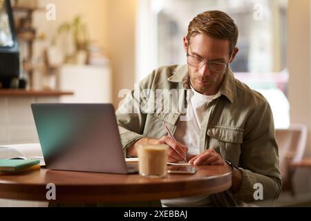 Image of focused young man in glasses, sitting in cafe, making notes, studying, attending online course, learning on remote from Stock Photo