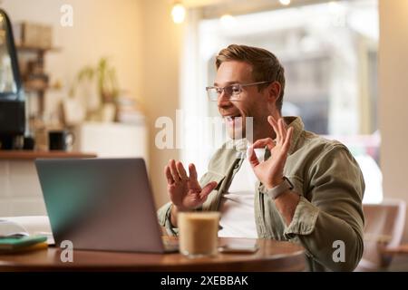 Portrait of young man, teacher, online tutor in glasses, showing okay, ok hand sign, giving lessons remotely from cafe, sitting Stock Photo