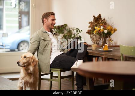 Portrait of handsome young man, cafe visitor, sitting with his dog, petting golden retriever and reading news on digital tablet, Stock Photo