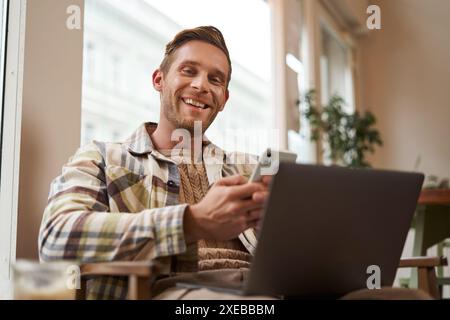Portrait of smiling, handsome young man relaxing in coffee shop, working remotely from cafe co-working space, using laptop and s Stock Photo
