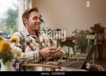 Image of handsome businessman working from cafe, drinking coffee and doing online project on his laptop, smiling and looking hap Stock Photo