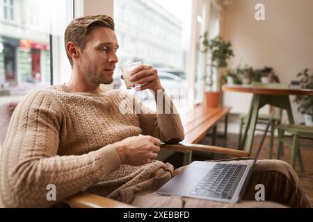 Working people and public space concept. Young handsome man, ux ui designer sitting with laptop in cafe, drinking cappuccino, lo Stock Photo