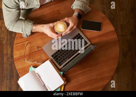 Top view. Male hands holding cup of coffee, man sitting at round table in a cafe, working on laptop, had his smartphone and note Stock Photo
