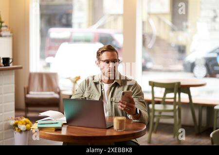 Lifestyle and working people concept. Young man in glasses sitting in empty cafe with laptop and notebook, checking his phone me Stock Photo
