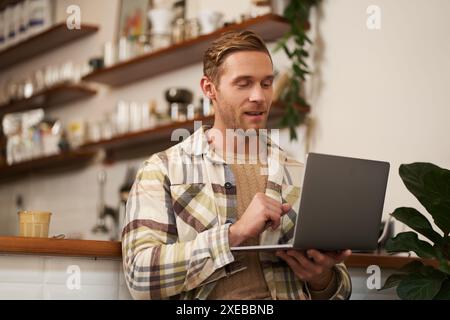 Portrait of handsome young man with laptop, sitting in cafe, looking at screen, doing work task, working as a freelancer, has re Stock Photo