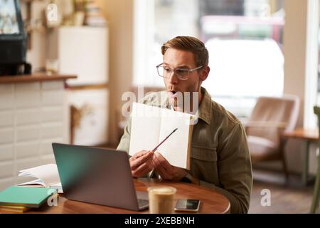 Portrait of handsome young man giving lessons online, showing exercises in his notebook, looking at laptop, recording video, sit Stock Photo